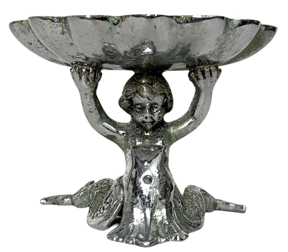 Saboneteira em metal prateado, em formato concha, sustentada por figura de putti (anjo) em relevo, com base decorativa. Peça de caráter ornamental, apresentando pátina e sinais do tempo. Mede 14cm x 10cm.