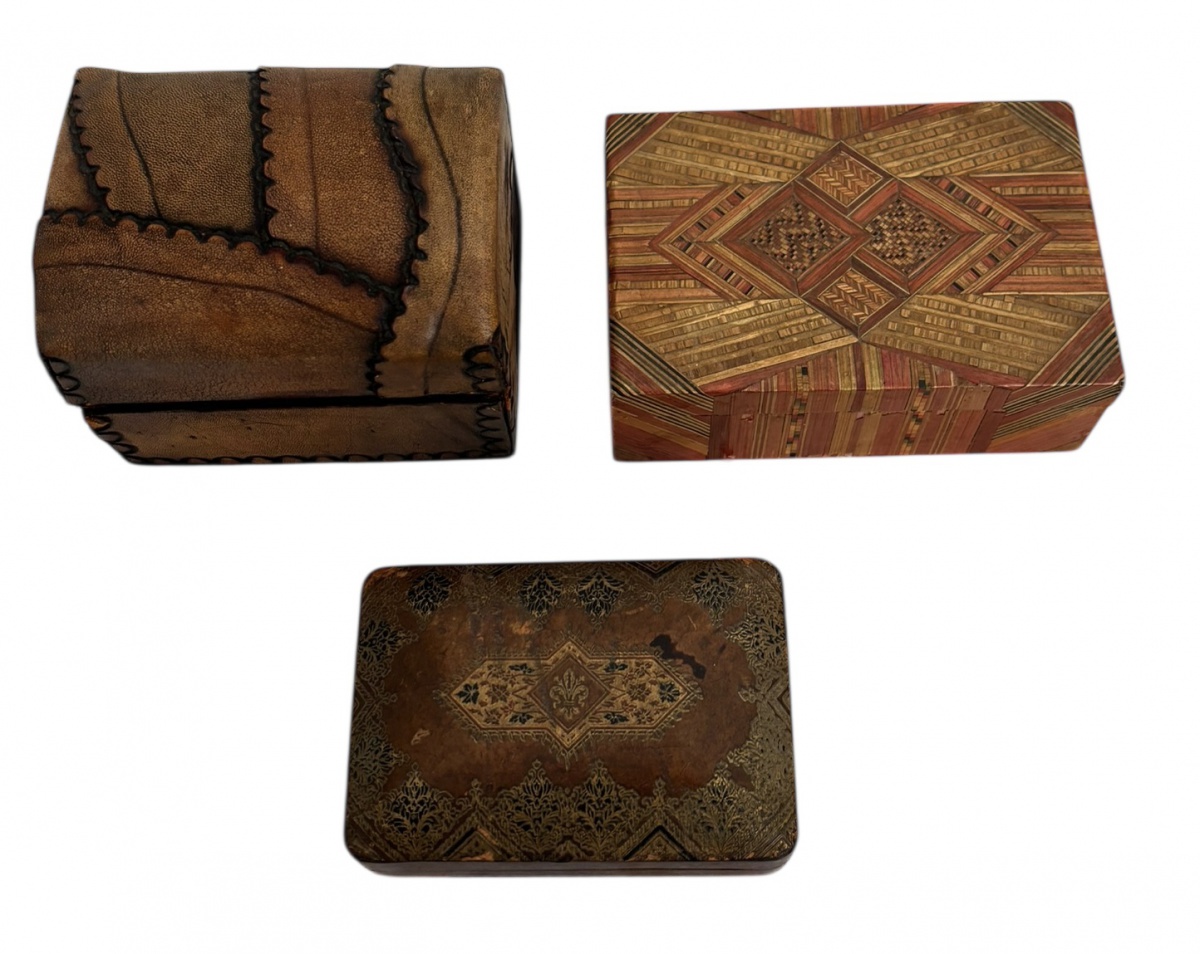 Lote composto por três caixas antigas decorativas, sendo: uma caixa revestida em couro com costuras aparentes e fecho metálico, uma caixa em marchetaria policromada com interior forrado em tecido vermelho, e uma caixa ...
