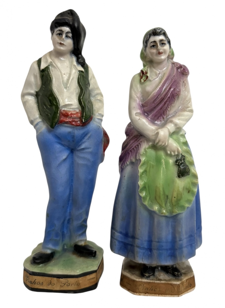 PORTUGAL - Par de antigas esculturas em porcelana europeia policromada, representando casal em trajes populares. Bases sextavadas com inscrição Vinhos do Porto e rótulos publicitários ao verso de vinícola. Peças ...
