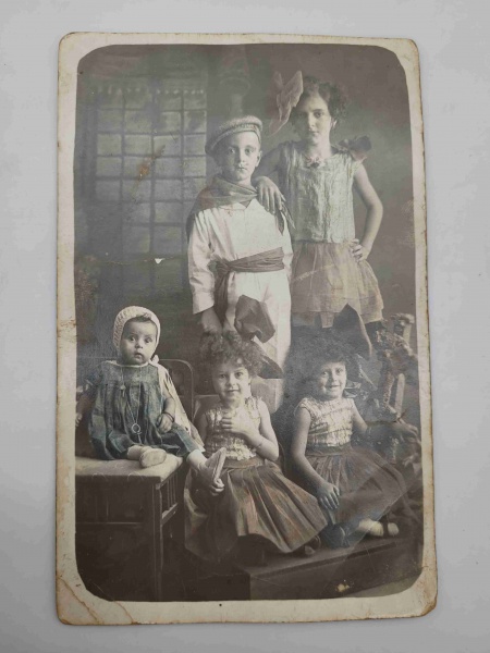 Antiga fotografia em formato de cartão postal retratando cinco crianças em trajes de época. Consta o ano de 1931 escrito manualmente no verso. Mede 13,5x8cm.