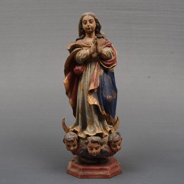 Brasil – sec. XIX. Imagem em madeira policromada e dourada representando Nossa Senhora da Conceição. Apresenta-se a Santa de pé, com o joelho direito em meia flexão em sua característica atitude de devoção com as mãos juntas em oração. Ricas vestes Túnica e manto em elegantes drapeados. Base formada por duas meias luas simbolizando que toda a luz da virgem é vinda e levada à Jesus cristo, seu filho em meio à nuvens três cabeças de querubins, revelando que a Virgem Maria está no céu, na glória de Deus, junto com os anjos. Alt. 39 cm.