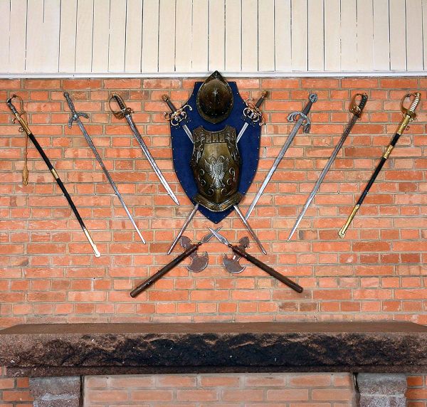 Conjunto decorativo composto por <b>armadura 8 espadas e 2 machadinhas</b>. Escudo central em metal repuxado com brasão em relevo e elmo superior. As peças apresentam lâminas metálicas polidas e punhos com empunhaduras ornamentadas.