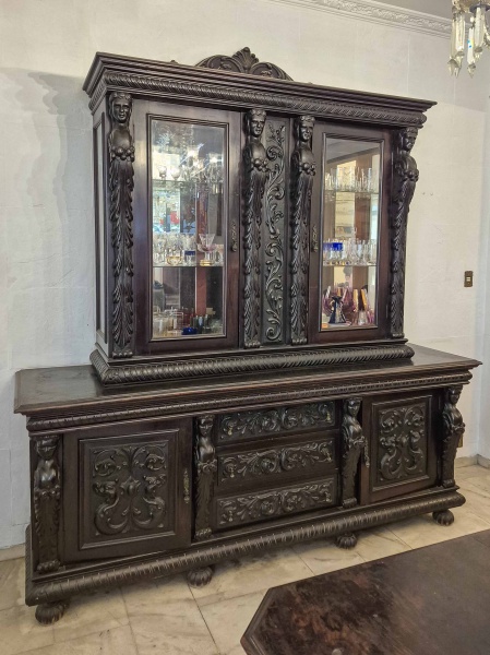 Antigo Móvel, composto por duas peças, em madeira nobre, com entalhes ornamentados com figuras femininas, conhecidas como Cariátides e fitomórficas, com características que remetem ao estilo manuelino e Renascentista, sendo a parte superior originalmente uma cristaleira de duas portas, com fundo espelhado e duas prateleiras em vidro, para armazenar Taças, usada sem os pés, sobre um buffet com duas portas e três gavetas. Dimensões: Totais: 240 cm X 242 cm X  66 cm; Buffet: 98 cm X 242 cm X 66 cm; 140 cm X 170 cm X 50 cm (Alt./Larg./Prof.). existem apenas três pés da cristaleiras, sendo que um falta o pino de encaixe, espelho com manchas do tempo. RETIRADA COM AGENDAMENTO PRÉVIO POR CONTA DO ADQUIRENTE OU TRANSPORTADORA INDICADA, EM CASA TÉRREA DA LAPA, RIO DE JANEIRO/RJ, IMPRETERIVELMENTE ATÉ O DIA 18.11, DATA DA ENTREGA DAS CHAVES DO IMÓVEL VENDIDO.Cariátides são esculturas femininas, usadas como colunas ou pilares arquitetônico, na antiga cidade grega de Cária, onde as mulheres eram conhecisas pela sua beleza e força. Foram usadas pela primeira vez na arquitetura grega clássica, cujo exemplo mais famoso são as cariátides do Erecteion, na Acrópole de Atenas. O uso de cariátides foi retomado posteriormente, como no movimento Renascentista, tornando-se populares em móveirs de decoração.