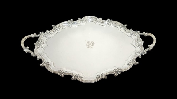 ELKINGTON & CO - Graciosa bandeja com alças em metal espessurado à prata com bordas onduladas, no centro ostenta monograma com as letras entrelaçadas "R", "G" e "O". Apresenta timbre da manufatura Elkington & CO na parte traseira da peça. Em ótimo estado. Medidas.: 63,5 x 42cm.