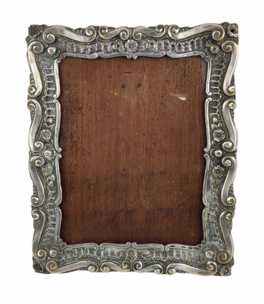 Porta-retratos de mesa em prata, decorado com trabalhos florais e volutas em relevo. Guarnições em madeira. Apresenta desgastes causados pelo tempo e pequenas mossas. Med. 33x27cm. Peso de prata 304g.