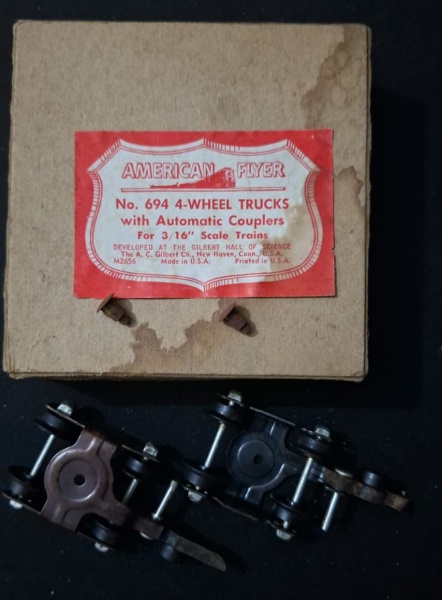 Conjunto de peças de trens de escala da marca American Flyer, especificamente o modelo No. 694 4-Wheel Trucks with Automatic Couplers, feito para trens de escala 3/16". As peças são dois caminhões (tr