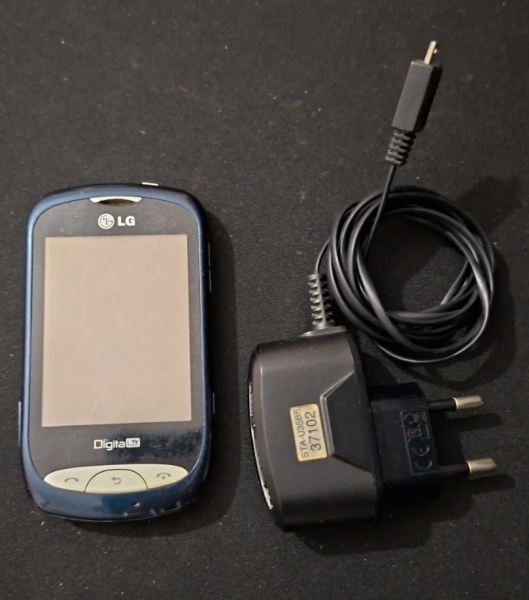 Aparelho de Celular com TV digital portátil LG* (modelo "Digital TV") acompanhado de seu carregador (modelo TA-J0585). Ele parece ser um dispositivo usado para assistir a transmissões de TV digital, p