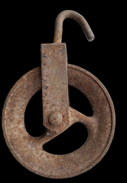 Roldana* (ou polia) de ferro fundido, com um gancho na parte superior para fixação e três rasgos no disco circular, típica de sistemas de elevação ou tração manual antigos. Diam 12cm