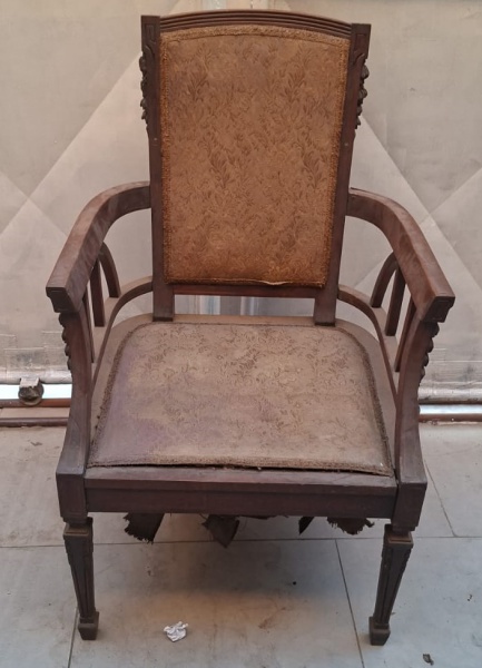 Cadeira de madeira antiga com braços, apresentando um estofado de tecido bege desgastado no assento e no encosto, com um padrão floral. A estrutura de madeira é escura e ornamentada, com detalhes escu