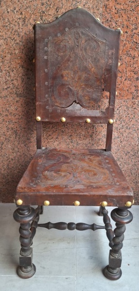Cadeira de madeira escura com detalhes entalhados, assento e encosto forrado com couro pirografado, encosto no estado, demanda reparo, apresentando um design ornamentado com motivos florais e volutas.