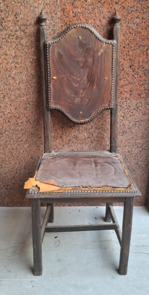 Cadeira antiga de madeira com um encosto alto e curvas ornamentadas no topo. O assento originalmente era revestido com um material tipo couro ou tecido marrom, mas está desgastado e rasgado, mostrando