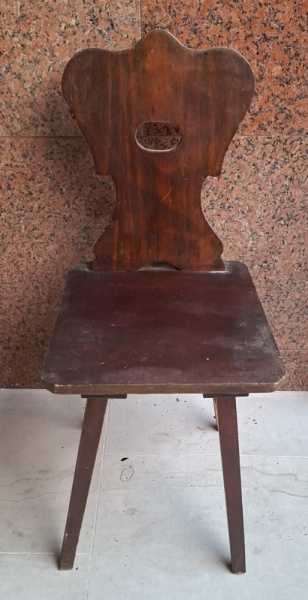 Cadeira de madeira escura com um encosto alto e recortado em forma oval no centro. O assento é quadrado e desgastado, e as pernas são retas e finas, também de madeira. A cadeira parece antiga e usada.