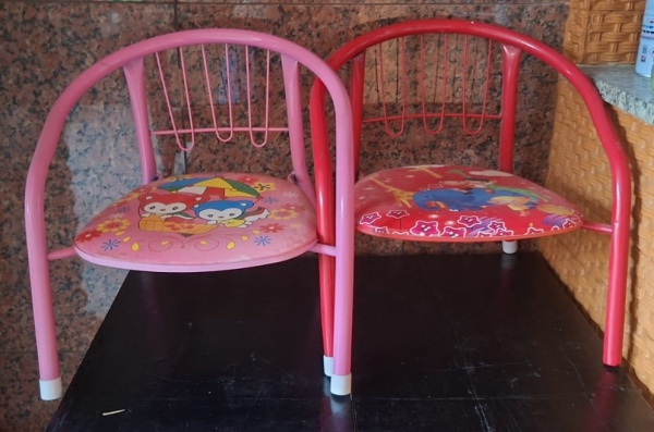 Par cadeira infantil vermelha com estrutura de metal e um assento almofadado também vermelho e uma rosa. O assento apresenta um desenho colorido de personagens infantis.