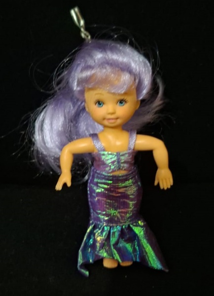 Boneca da coleção Paula, com cabelo azul longo e olhos azuis. Ela está vestindo um vestido verde/teal brilhante em formato de sereia. A boneca está embalada em um tubo cilíndrico transparente com uma