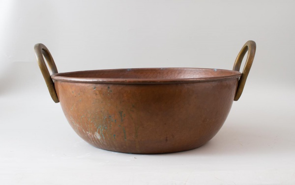 Tacho em cobre martelado com alças laterais em metal dourado, peça de formato circular utilizada para decoração ou preparo culinário. Apresenta marcas do tempo, oxidação, desgastes e sinais de uso con