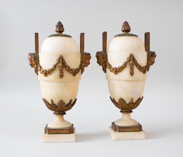 Par de urnas decorativas em alabastro com montagens em metal patinado, tampas removíveis encimadas por pinhas e aplicações ornamentais em guirlandas e mascarões laterais. Peças em estilo clássico com