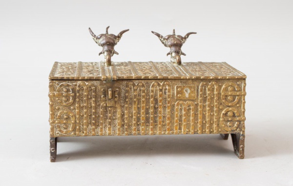 Caixa decorativa em metal dourado, ricamente trabalhada com inscrições e ornamentos em relevo, tampa com aplicações figurativas de cabeças bovinas, apoiada sobre pés, peça de forte influência oriental