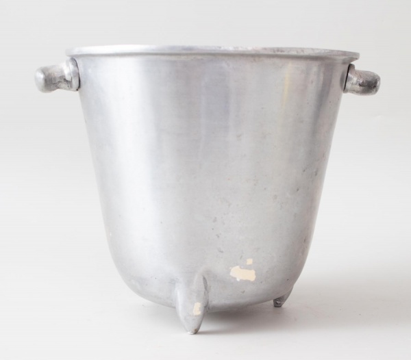 Balde para gelo em metal prateado, corpo troncocônico com borda reforçada, dois pinos laterais para encaixe de alça e três pés pequenos na base. Acabamento escovado com pátina de uso. Peça funcional c