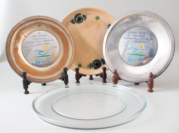 Lote contendo dois pratos comemorativos em metal alusivos aos 25 anos do Atlântico Sul e um prato decorativo em cerâmica esmaltada com motivos abstratos; medindo os maiores 29 cm de diâmetro; apresent