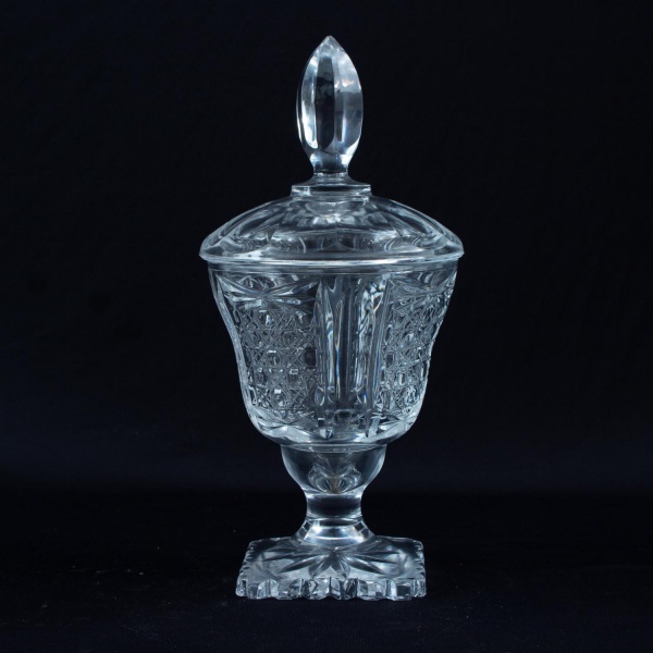 Bomboniere em cristal lapidado com tampa e pegador facetado, apoiada sobre base quadrada trabalhada, medindo 26 cm de altura; apresenta sinais de uso, marcas do tempo e pequenos desgastes.