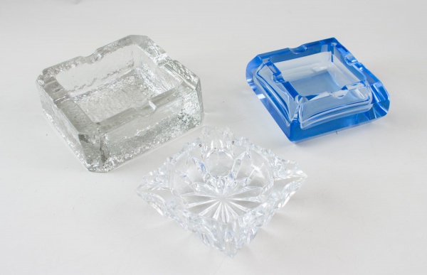 Lote contendo três cinzeiros, sendo dois em cristal incolor e um em vidro azul, de formatos quadrados variados; apresentam sinais de uso, marcas do tempo e pequenos desgastes.