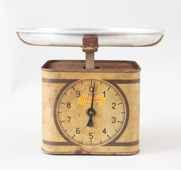 Balança doméstica antiga de mesa, com prato metálico superior e mostrador analógico frontal, marca London; apresenta marcas do tempo, oxidação, desgastes e sinais de uso.