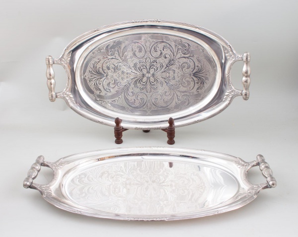 Par de bandejas ovais em metal prateado com alças integradas, medindo 54 cm de comprimento cada; uma delas com fundo ricamente gravado em motivos ornamentais; apresentam sinais de uso, marcas do tempo