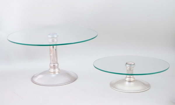 Par de boleiras Riva com tampos circulares em vidro e bases em metal prateado, medindo a maior 19 cm de altura e 37 cm de diâmetro; apresentam sinais de uso, marcas do tempo e pequenos desgastes.