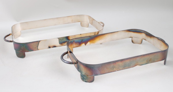 Par de suportes para rechaud em metal com alças laterais, formato quadrado; apresentam sinais de uso, marcas do tempo, oxidação superficial e pequenos desgastes.