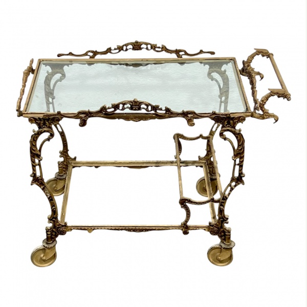 Carrinho de chá/bar em metal trabalhado com decoração vazada, rodízios e tampo de vidro, medindo 57 cm de altura e 73 x 43 cm; apresenta marcas do tempo, desgastes, ausência de um dos vidros e vidro r