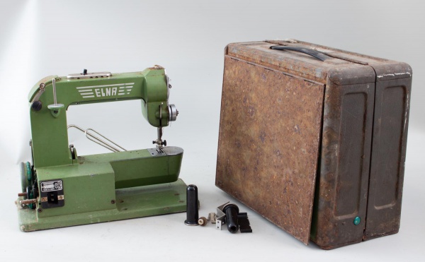 Máquina de costura portátil Elna, fabricada na Suíça, acondicionada em estojo metálico original, medindo aproximadamente 30 cm de altura e 35 cm de comprimento; apresenta oxidação acentuada no estojo,