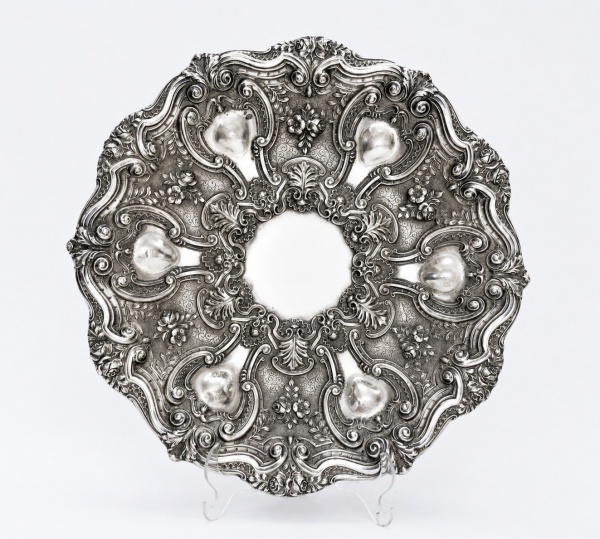 Medalhão em prata teor 833, ricamente trabalhado em relevo com decoração em estilo rococó, apresentando volutas, elementos florais e reservas em formato de cartelas distribuídas ao redor do centro, me