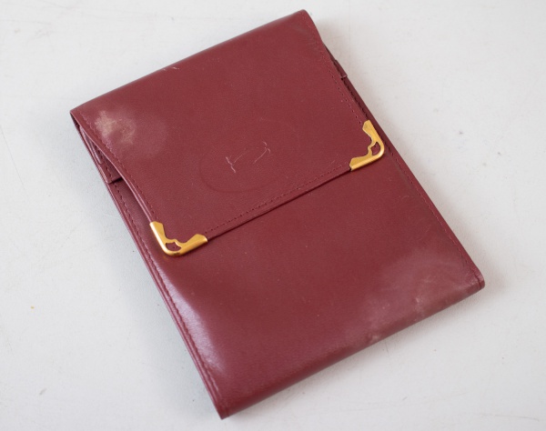 Carteira de bolso Must de Cartier em couro na cor bordeaux, com acabamento liso, logotipo da grife em relevo na face frontal e detalhes de cantoneiras em metal dourado, interior com inscrição Cartier