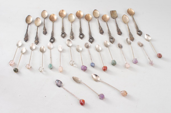 Lote composto por 25 colheres de café em metal, com modelos e acabamentos variados, incluindo exemplares com cabos ornamentados e cabos decorados com aplicações coloridas. Maior peça medindo 13 cm de