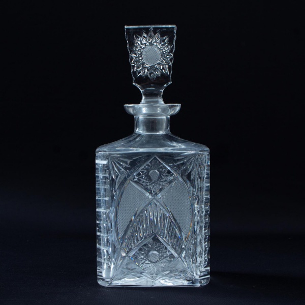 Garrafa licoreira em cristal lapidado, com corpo retangular de ombros inclinados e tampa original lapidada, ricamente decorada com padrões geométricos e motivos estrelados, medindo 28 cm de altura, co