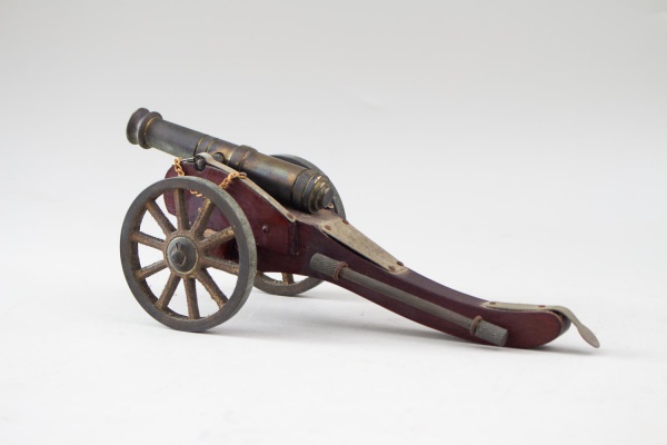 Canhão decorativo em metal e madeira, com detalhes em latão, rodas raiadas e vareta de limpeza fixada à carreta, medindo 10 cm de altura e 28 cm de comprimento, apresenta marcas do tempo, sinais de ox