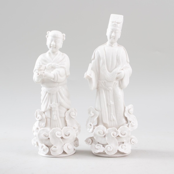 Par de esculturas orientais em porcelana branca, representando figuras tradicionais sobre base com nuvens estilizadas em relevo, medindo 23 cm de altura cada, com marcas do tempo.