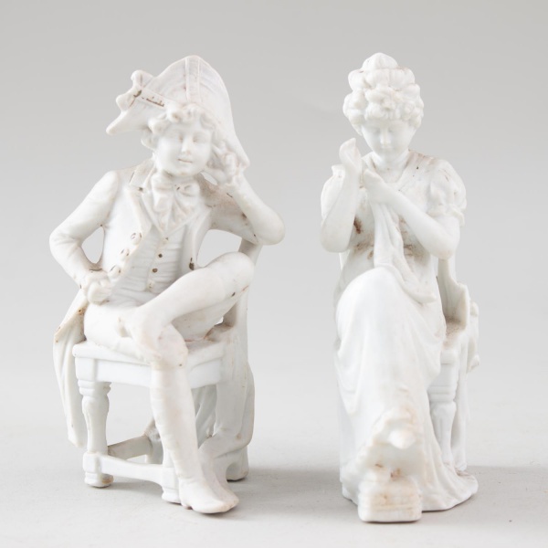 Escultura em porcelana branca representando casal em cena galante, com figuras sentadas em cadeiras e trajes de época, medindo 12 cm de altura, apresentando marcas do tempo e desgastes compatíveis com