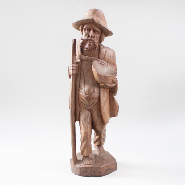 Escultura representando mendigo em madeira entalhada, com chapéu, cajado, cuia e cantil às costas, sobre base integrada; altura: 41 cm; apresenta marcas do tempo e desgastes naturais.
