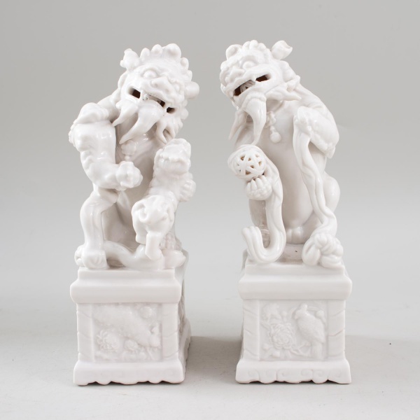 Par de esculturas orientais representando cães de Fó (Foo Dogs) em porcelana branca vitrificada, sobre bases decoradas em relevo; altura: 23 cm cada; produzidas na China; apresentam marcas do tempo e