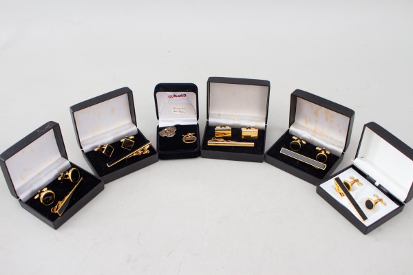 Lotão com 6 conjuntos de abotoaduras e prendedores de gravata, em metal dourado, com diferentes modelos e detalhes em esmalte preto, acompanhando estojos. Inclui um conjunto temático Cunard (Diamond &