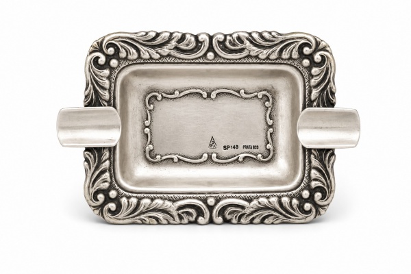 Cinzeiro em prata teor 833, de formato retangular com apoios laterais para charuto, borda ricamente ornamentada em relevo com motivos vegetalistas de inspiração rococó, medindo 10 cm de comprimento e
