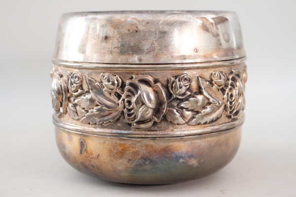 Cachepot em metal espessurado a prata, de corpo cilíndrico com faixa decorada em relevo de rosas, medindo 20 cm de altura e 21 cm de diâmetro, apresentando marcas de uso, oxidação e marcas do tempo.