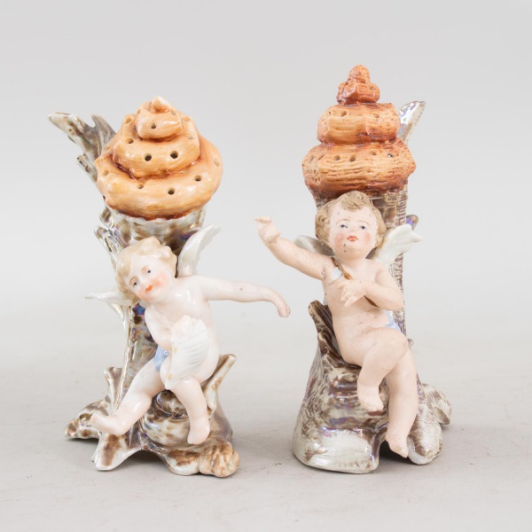 Par de paliteiros antigos em porcelana de origem europeia, marca Otto, modelados em forma de querubins apoiados sobre estruturas em formato de conchas, com reservatórios perfurados, medindo 14 cm de a