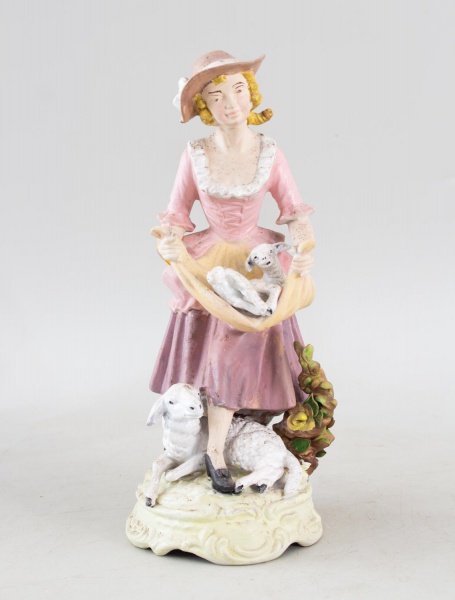 Escultura em porcelana alemã, ao estilo Capodimonte, representando camponesa trajando vestes típicas e carregando carneirinho em tecido, com outro carneiro aos seus pés, ricamente policromada e marcad