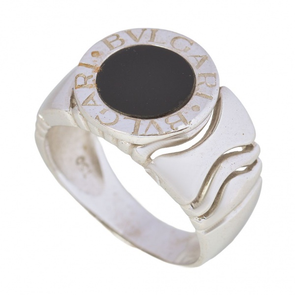 Anel Bvlgari em ouro branco 18K com ônix central, peso: 7,1 g, aro: 15. Modelo de design robusto e elegante, com aro espesso e face redonda destacando a inscrição BVLGARI BVLGARI. Peça unissex de esti