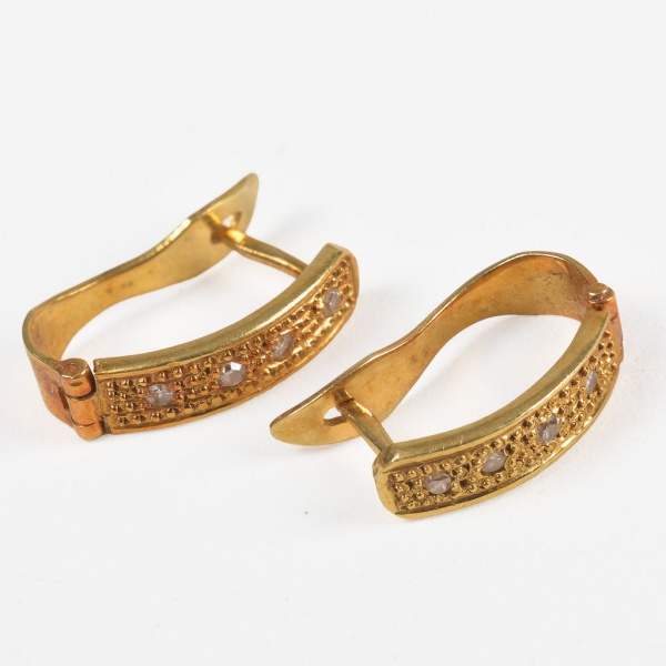 Par de brincos em ouro amarelo 18K, peso total: 3,1 g. Modelo delicado com frente levemente curvada, cravejada com pequenas pedras claras, possivelmente brilhantes. Fecho tipo alavanca, garantindo seg