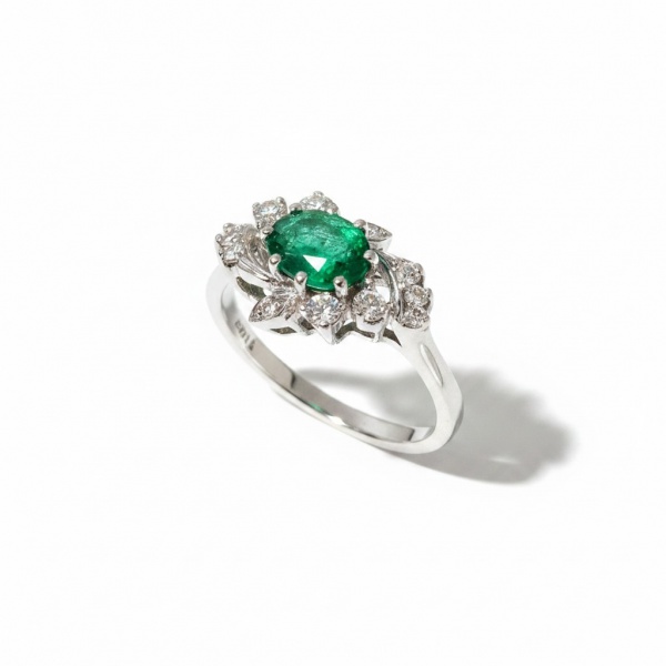 Anel em ouro branco 18k (750), pesando aproximadamente 4,93 g. Apresenta esmeralda natural central em lapidação oval com peso aproximado de 0,65 ct, em verde vivo e translúcido, circundada por brilhan