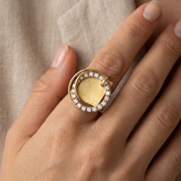 Anel de design contemporâneo em ouro amarelo 18k, peso 15,9g. Apresenta volumoso elemento circular central com disco em ouro de superfície escovada/texturizada, circundado por aro cravejado com diaman