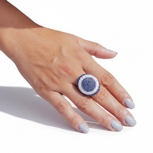 Anel cocktail em ouro branco 750, peso 13,08g, aro 18,5. Design circular domed inteiramente pavimentado com mais de 150 pontos de safiras azuis e 60 pontos de diamantes em halo radiante.
